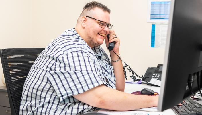 A man in an office setting is looking at the computer and on the phone. He is smiling.