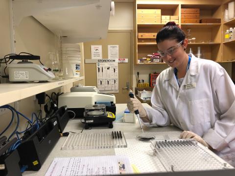 Rachel in lab coat and glasses, holding a dropper above a sample in a laboratory setting