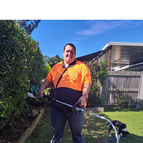 Nicholas proudly standing in a garden with a gardening tool and a dog visible in the background