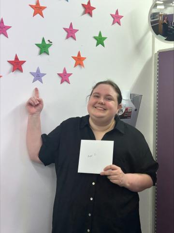 An individual wearing black clothing standing in front of a white wall with colourful stars on it. She is holding a white piece of paper up, similar to a certificate.